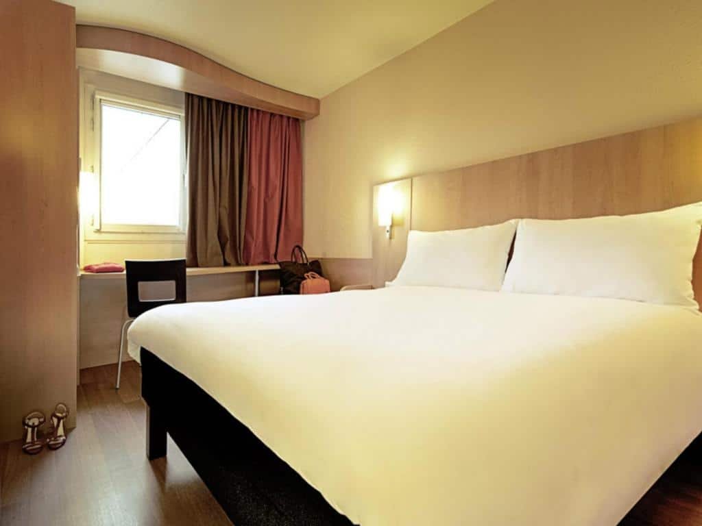 Quarto do ibis Porto Centro São Bento com cama de casal do lado direito e do lado esquerdo da cama uma mesa de trabalho. Representa hotéis ibis no Porto.