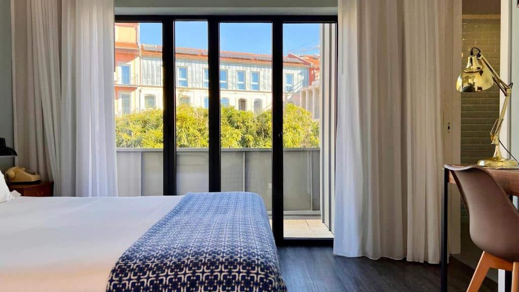 Quarto do Jardim da Batalha Boutique Hotel by Shiadu com cama de casal do lado esquerdo da imagem, uma mesa de trabalho com cadeira em frente a cama e do lado esquerdo uma porta de vidro que dá acesso a varanda. Representa hotéis no centro do Porto.
