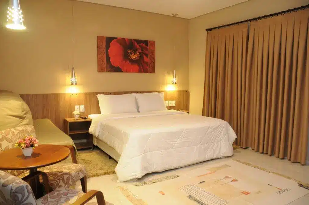 Quarto novo no O Recanto Pousada com uma cama de casal, um sofá, uma mesinha com duas poltronas, uma varanda com cortinas, tudo decorado em tons de branco, bege e madeira.