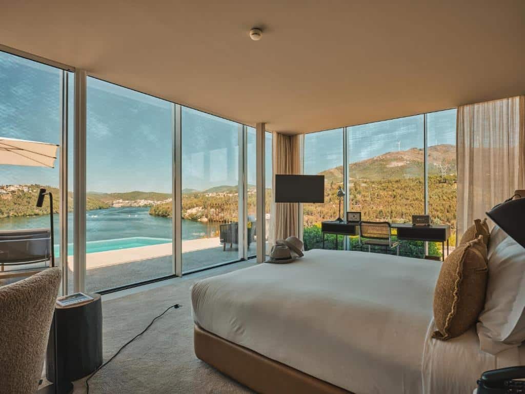 Quarto do Octant Douro com cama de casal do lado direito da imagem, do lado esquerdo da cama uma mesa de trabalho com cadeira, em frente a cama porta de vidro com vista para a a piscina e ao fundo o rio Douro. Representa hotéis do Vale do Douro.