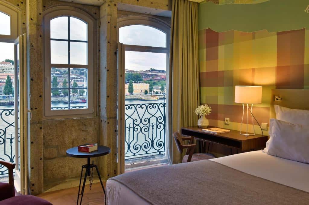 Quarto do Pestana Vintage Porto Hotel & World Heritage Site com cama de casal do lado direito da imagem, do lado esquerdo da cama uma mesa com cadeira ao lado de uma porta que dá acesso a varanda com vista para o rio. Representa hotéis com vista do Douro.