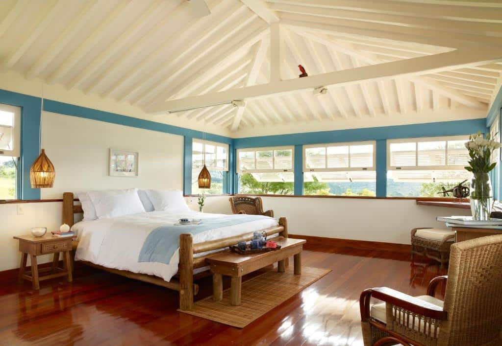 Hospedagem no Lake Vilas Charm Hotel. Um quarto grande de luxo, com uma cama de casal grande, uma cadeira do lado, duas cadeiras de frente e entre as cadeiras uma televisão. O quarto tem janelas grandes ao redor.