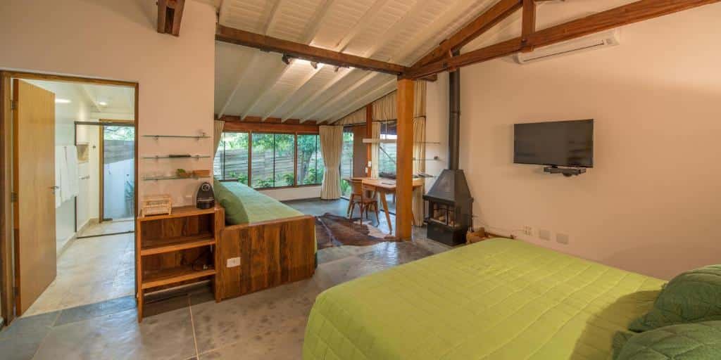Uma cama de casal no canto direito, na parede uma televisão e uma lareira, na Fazenda Capoava. No canto esquerdo a vista do banheiro. De frente para a cama um sofá, uma mesa de escritório e a vista da mata.