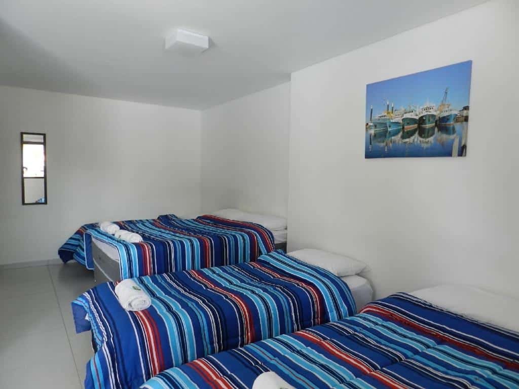 Quarto da Pousada Acqua Bella, uma cama de casal e duas de solteiro, com toalhas, travesseiros e cobertas sob a cama, e um quadro com barcos desenhados preso na parede, para representar pousadas em Riviera de São Lourenço