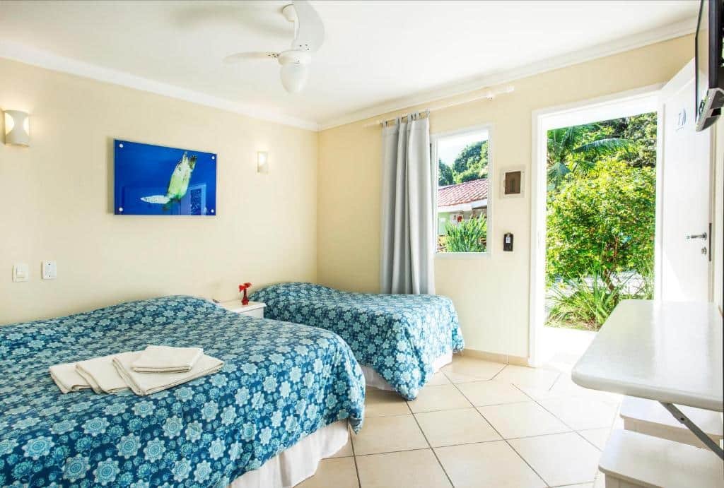 Quarto na Pousada Azul da Cor do Mar com uma cama de casal e uma de solteiro, há uma televisão, uma bancada com dois banquinhos e uma janela com vista para o jardim, para representar pousadas pé na areia em Maresias