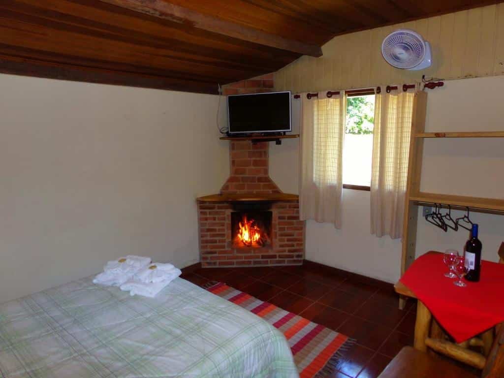 Quarto com cama de casal, lareira, TV, ventilador e uma mesa com duas taças e garrafa de vinho.