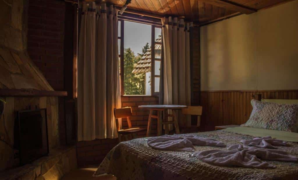 Quarto com cama de casal, mesa com duas cadeiras ao lado de uma janela com vista para fora e uma lareira.
