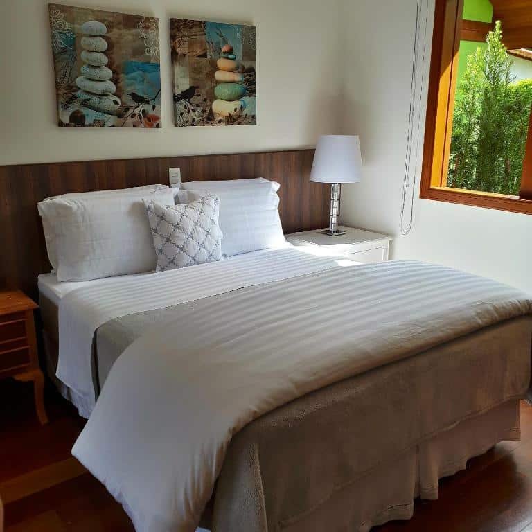 Quarto com cama de casal, quadro na parede, abajur branco ao lado e uma janela com vista para fora.