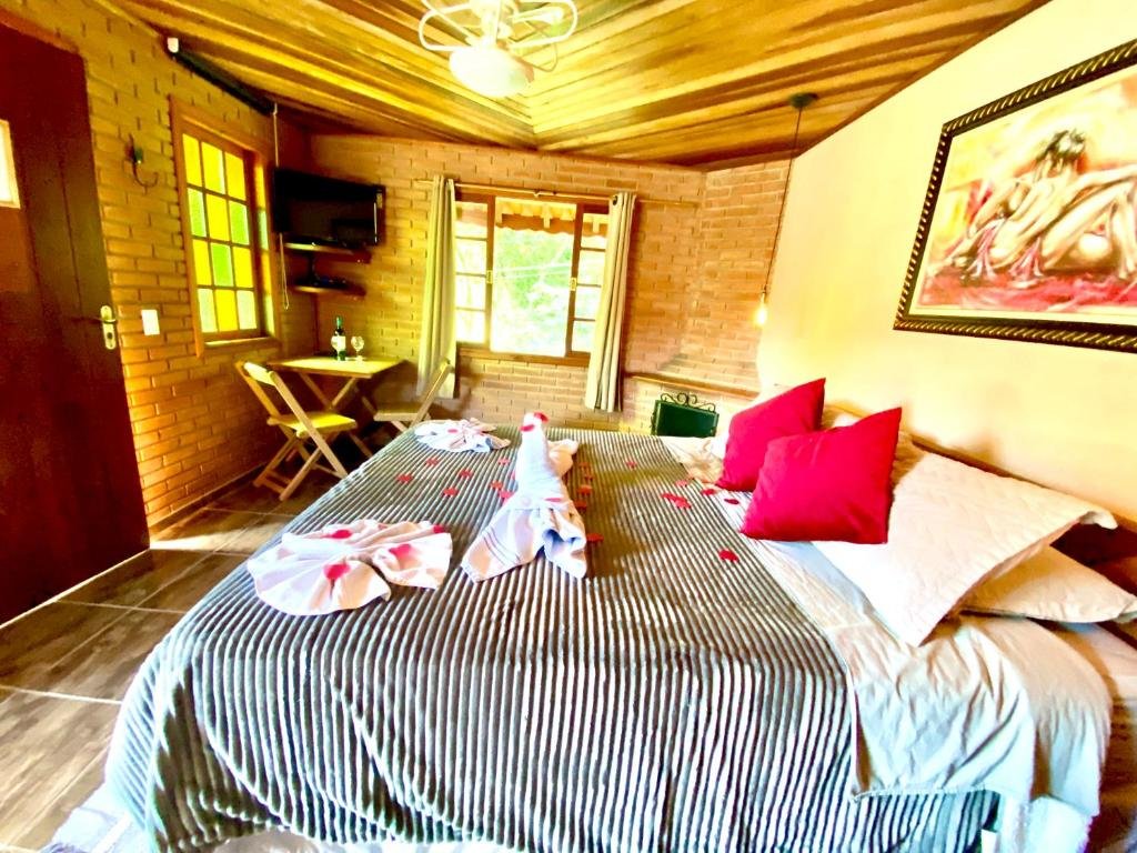 Quarto com cama de casal, quadro na parede, lareira, janela de vidro, TV na parede e logo em baixo uma mesa com duas cadeiras.