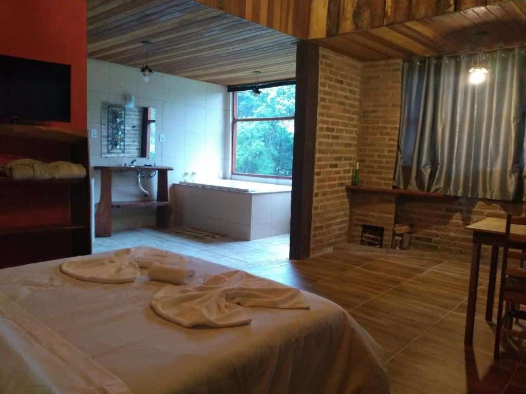 Quarto com cama branca e duas toalhas em cima e banheira ao fundo perto da janela de vidro.