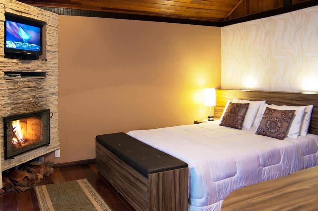 Cama de casal, com abajur ao lado, TV na parede e lareira.