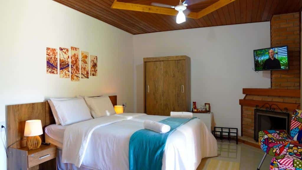 Quarto com cama de casal, guarda-roupa, lareira, poltronas, abajur e uma TV.