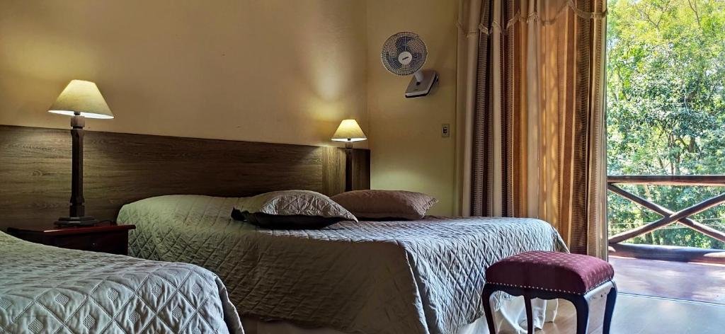 Quarto da pousada taverna do golf em Teresópolis com duas camas, abajur, ventilador na parede e sacada com vista para as árvores, ilustrando post pousadas em Teresópolis.