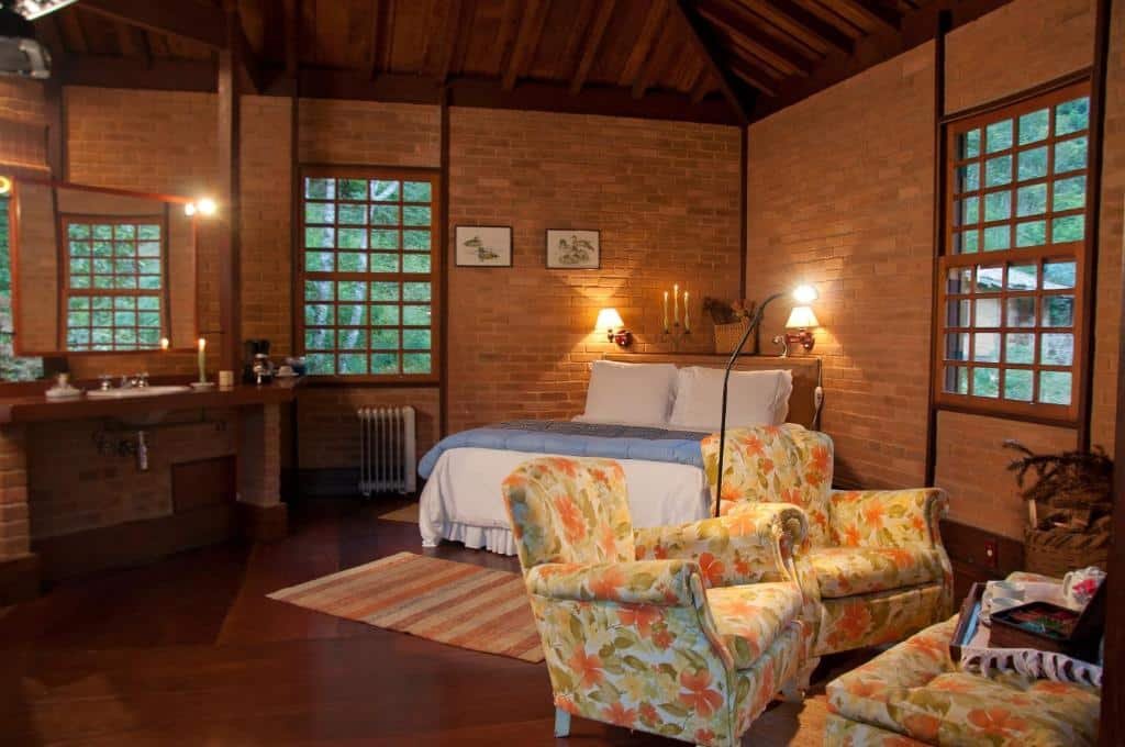 Quarto com paredes de tijolos, cama de casal, poltronas coloridas e janelas de madeira e vidro.