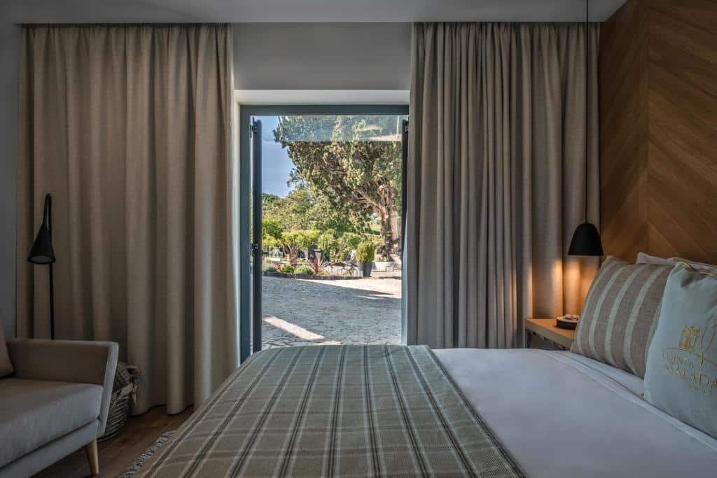 Quarto do Quinta da Salada – Turismo Rural com cama de casal do lado direito da imagem, em frente a cama uma poltrona e do lado esquerdo da cama uma cômoda com luminária e do lado da cômoda uma porta aberta que dá acesso ao térreo.