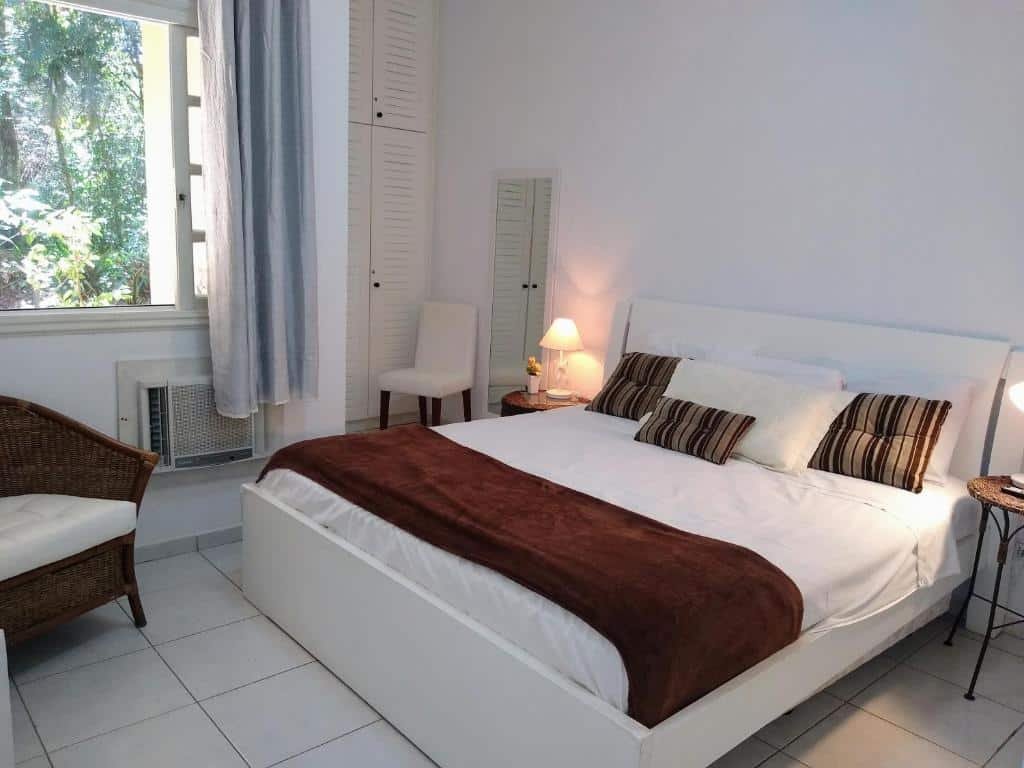 Quarto em Riviera Flat Service- 109, uma cama de casal, um armário, uma cadeira, uma mesinha de cabeceira com abajur, um ar-condicionado, uma poltrona e uma janela com cortinas, para representar pousadas em Riviera de São Lourenço