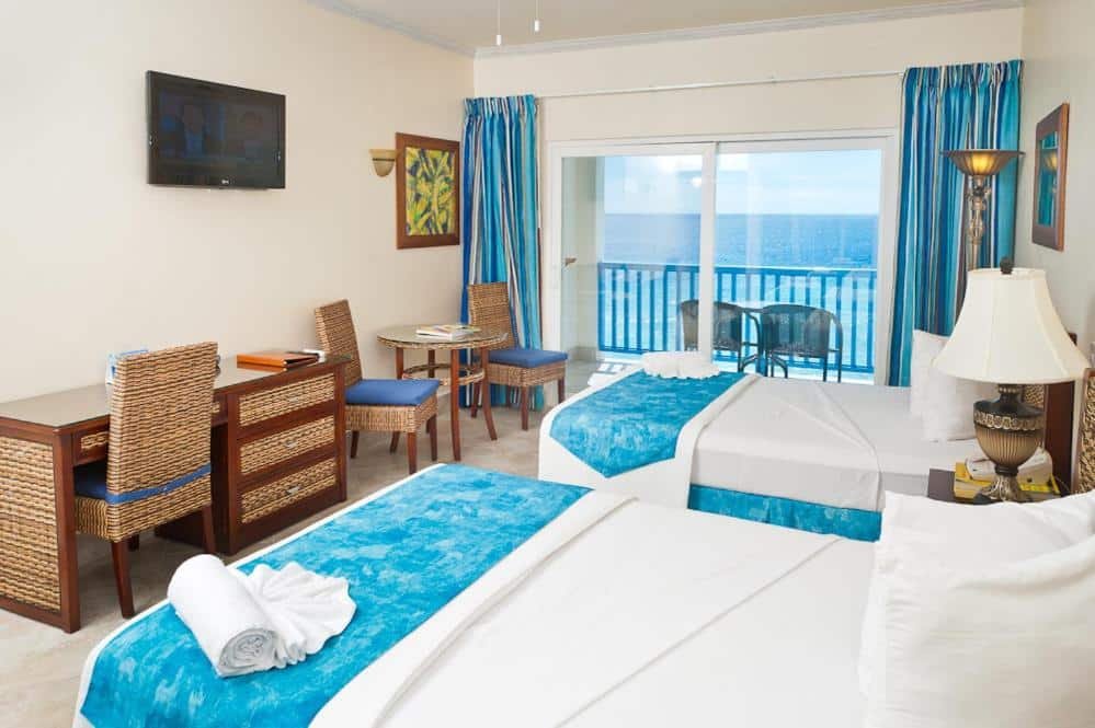 Quarto do Rostrevor Hotel. Duas camas de casal do lado direito, entre elas uma cômoda e um abajur. Do lado esquerdo uma mesa de trabalho, uma mesa com duas cadeiras e uma TV. No fundo a porta de vidro da varanda com acesso a varada e vista do mar.