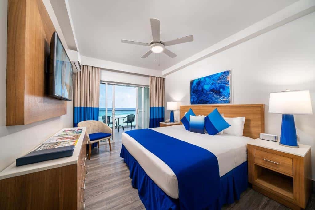 Quarto do South Gap Hotel que mostra do lado direito uma cama de casal com travesseiros e cobertas em cima e de cada lado da cama tem uma mesa de cabeceira. Em frente a cama tem uma estante e um painel de madeira com uma tv. Já do lado direito do quarto tem uma poltrona e uma porta de vidro com cortinas que dá acesso a varanda do quarto.