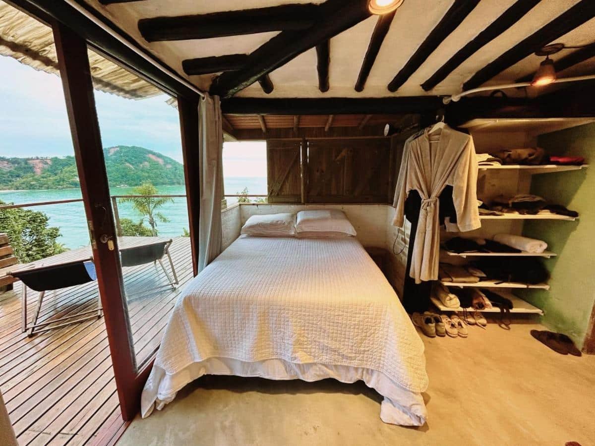 Quarto da Suíte e sala - Vista Barra do Sahy com cama de casal no centro do ambiente do lado esquerdo da cama um roupão pendurado e várias prateleiras com roupa e do lado direito da cama uma porta de vidro que dá acesso a varanda.
