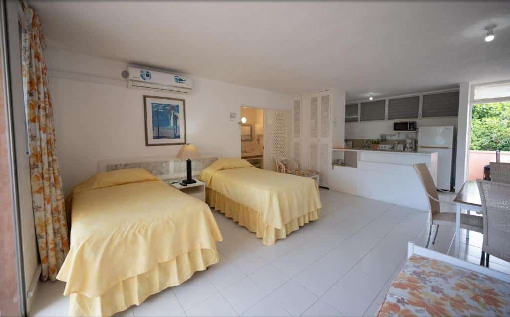 Quarto do The Monteray Hotel. Do lado direito uma mesa com cadeiras e uma cama de solteiro. Do lado esquerdo duas camas de solteiro, entre elas uma cômoda com abajur e um ar-condicionado. No fundo a cozinha.