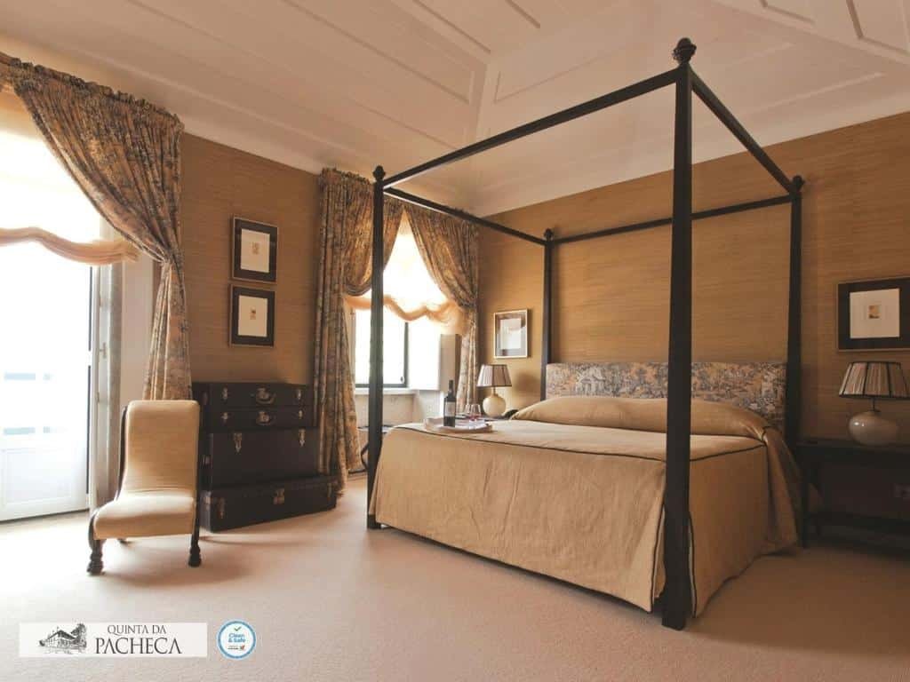 Quarto do The Wine House Hotel – Quinta da Pacheca com cama de casal do lado direito, com duas cômodas com luminárias ao lado e do lado esquerdo da cama em frente a cama uma poltrona. Representa hotéis no Vale do Douro.