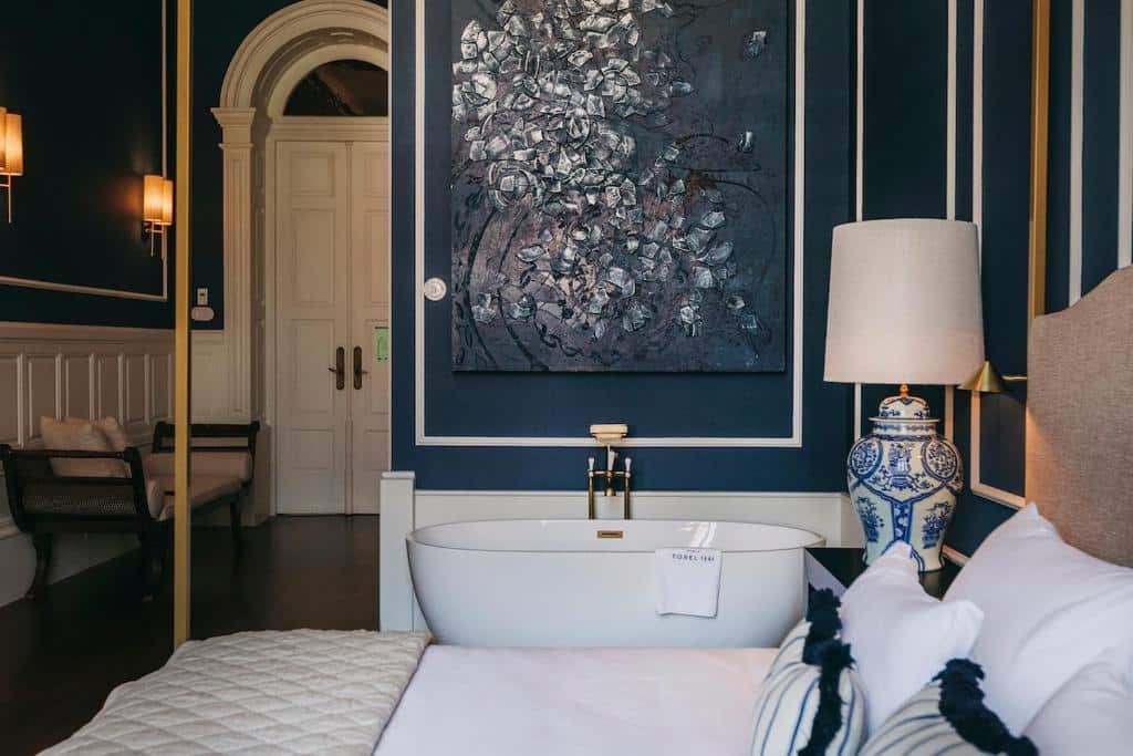 Quarto do Torel 1884 Suites & Apartments com cama de casal do lado direito, uma cômoda ao lado cama com luminária e ao lado uma banheira de hidromassagem. Representa hotéis no Porto.