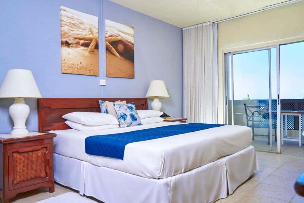 Quarto do Tropical Sunset Beach que mostra uma cama de casal do lado esquerdo e de cada lado da cama tem uma mesa de cabeceira com um abajur. Já do lado esquerdo da cama tem uma porta de vidro com cortinas que dá acesso a varanda do quarto.