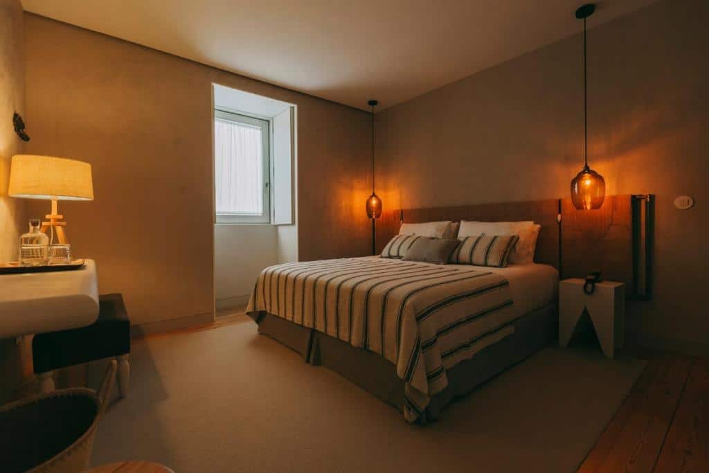 Quarto do Ventozelo Hotel & Quinta com cama de casal do lado direito da imagem com duas luminárias compridas presa no teto que fica em cada lado da cama e em frente a cama uma mesa de trabalho com banco estofado. Representa hotéis no Vale do Douro.