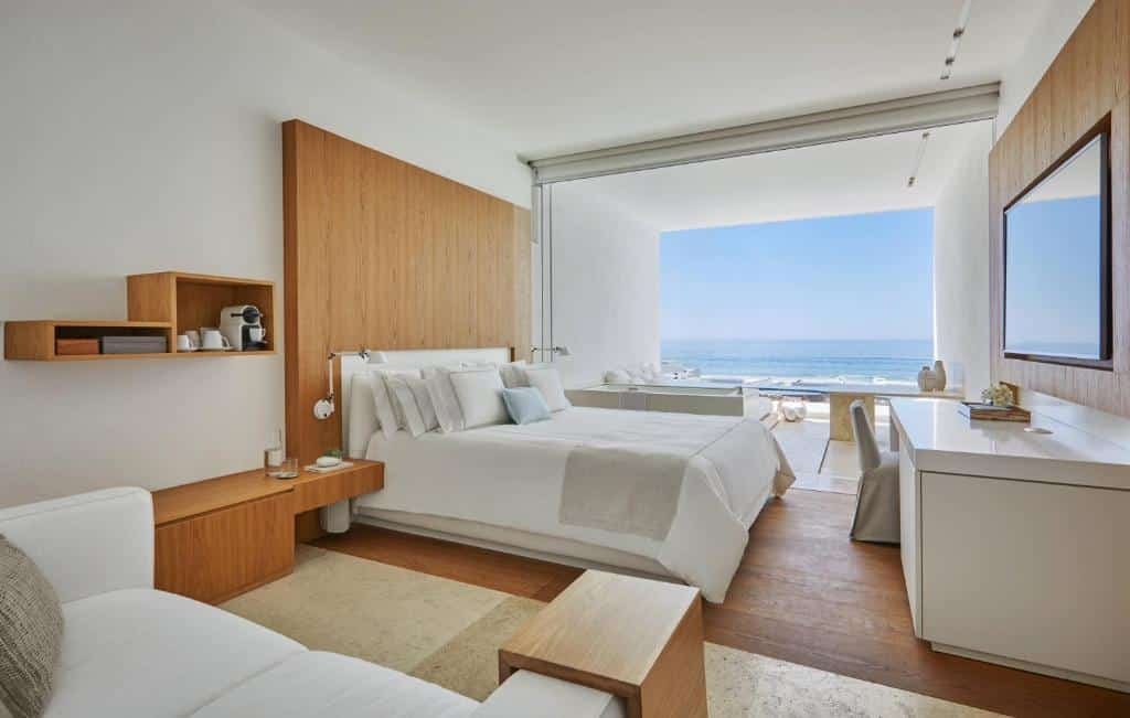 Quarto do Viceroy Los Cabos com cama de casal, duas cômodas com luminárias, TV em frente a cama com mesa de trabalho. Sofá do lado direito e do lado esquerdo com janela ampla em com vista para o mar.