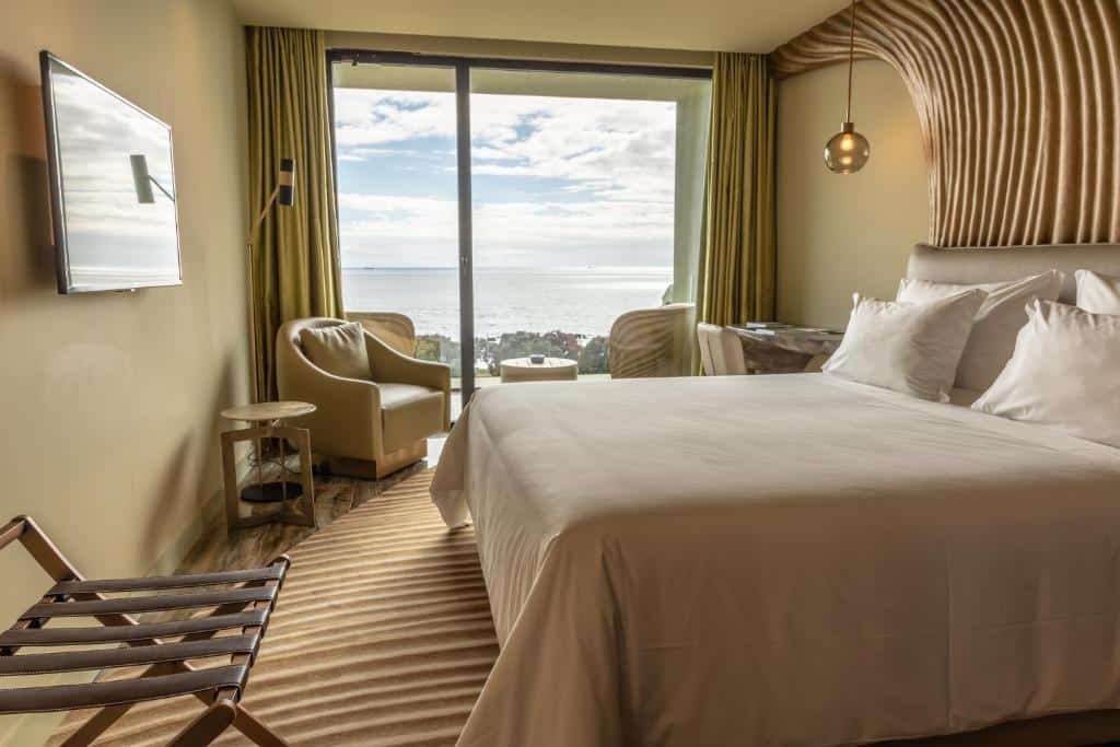 Quarto do Vila Foz Hotel & SPA – member of Design Hotels  com cama de casal do lado direito da imagem em frente a cama uma TV presa na parede do lado esquerdo do quarto uma poltrona e logo ao fundo uma varanda com sacada. Representa melhores hotéis no Porto.