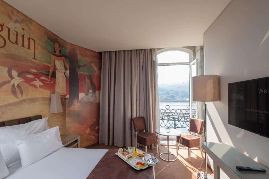 Quarto do Vila Gale Porto Ribeira com cama do lado esquerdo, em frente a cama uma cômoda com TV e do lado esquerdo uma mesa com duas cadeiras perto de uma porta que dá acesso a varanda. Representa hotéis com vista do Douro.