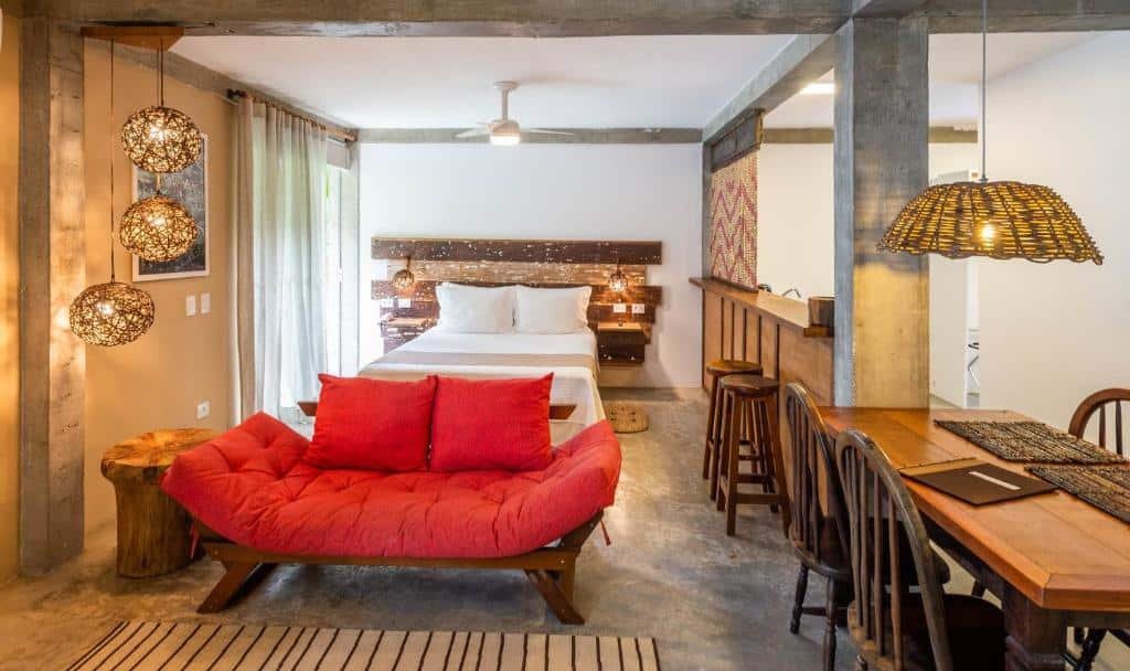 Quarto com dois ambientes na Villa Sapê Pousada com uma cama de casal e uma sacada ao lado esquerdo, mais a frente há um sofá de dois lugares e uma mesa com quatro cadeiras do lado direito, tudo decorado de forma moderna com madeira rústica, para representar resorts em Ubatuba