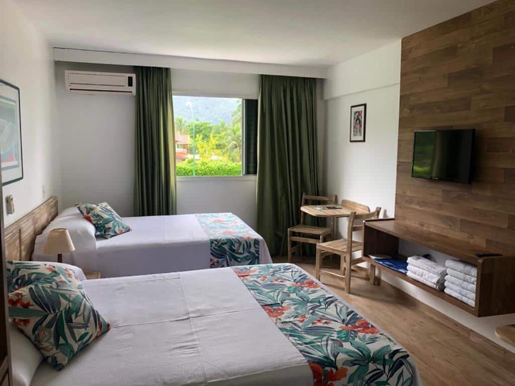Quarto no Wembley Inn Hotel com duas camas de casal, uma janela com cortinas, um rack com televisão, uma pequena mesa com duas cadeiras, tudo decorado em madeira e tons de verde, para representar resorts em Ubatuba