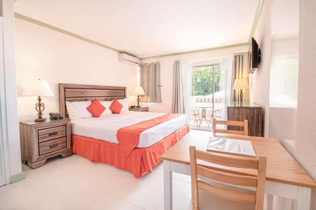 Quarto do Worthing Court Apartment Hotel que mostra do lado esquerdo uma cama de casal e de cada lado da cama tem uma mesa de cabeceira com um abajur. Em frente a cama tem uma mesa com duas cadeiras, um móvel de madeira e uma tv na parede. No quarto também tem uma porta de vidro com cortinas que dá acesso a varanda do quarto.
