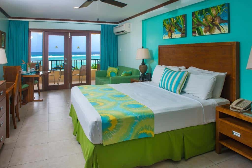 Quarto do Yellow Bird Hotel que mostra uma cama de casal do lado direito do quarto, de cada lado da cama tem uma mesa de cabeceira e dois quadros na parede. Em frente a cama tem um móvel de madeira e do lado uma mesa de madeira com cadeira. No quarto tem também um sofá e do lado do sofá tem uma porta de vidro com detalhes em madeira e cortinas que dá acesso a varanda do quarto com vista para o mar.