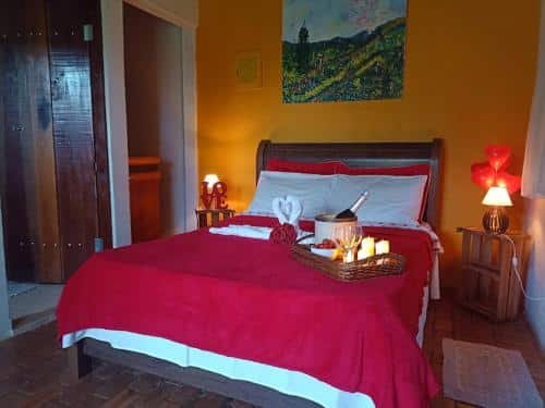 Quarto do Recanto Cheio de Cheiro, com uma cama de casal com lençol vermelho, bandeja com velas, balde de champanhe e toalhas enroladas em cima. Há uma parede laranja, um quadra com desenho de natureza em cima, e uma escrivaninha ao lado com luminária de corações acesa. No lado esquerdo tem uma porta de madeira