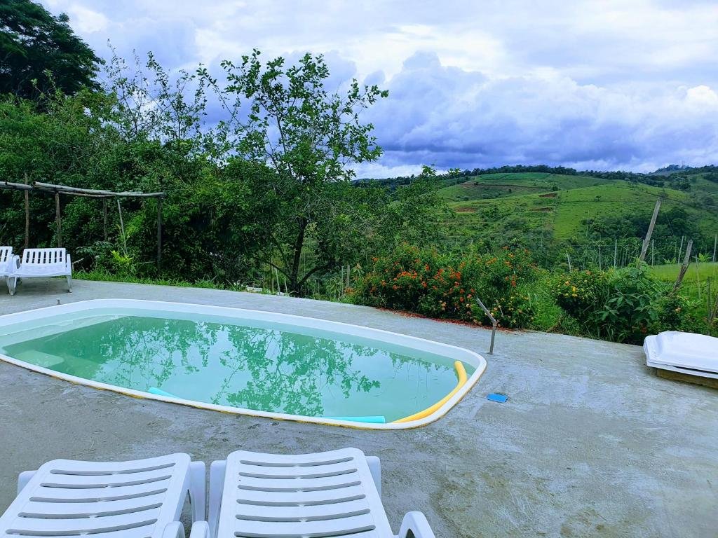Piscina da Recanto das Acácias, uma das pousadas em Guararema, com espreguiçadeiras brancas em volta e vista para montanhas e bastante natureza no lado direito.
