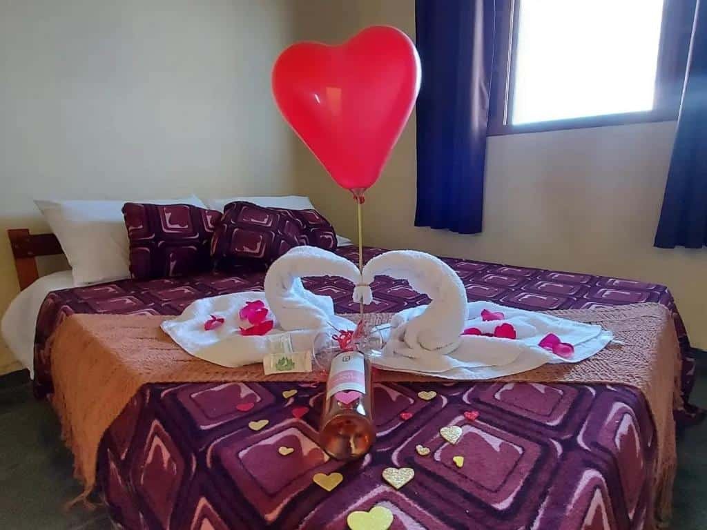 Cama do quarto da Recanto Varandas Guararema, com duas toalhas enroladas formando duas esculturas de cisnes, pétalas vermelhas e um balão de coração vermelho em cima. No lado direito tem uma janela aberta, com uma cortina marrom