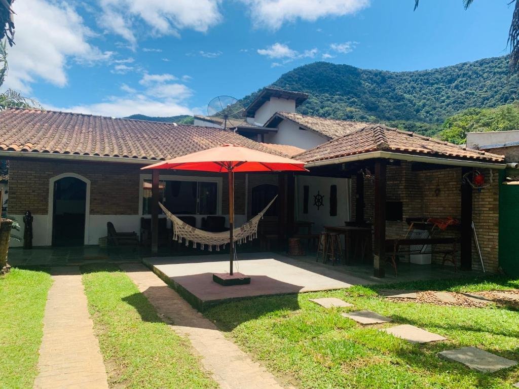 Quintal e varanda da casa de temporada "Refugio Maresias". O quintal é de gramado e possui caminhos de concreto. Ao fundo está a casa. Na parte da varanda há uma rede, um guarda-sol, mesas, cadeiras e uma churrasqueira.