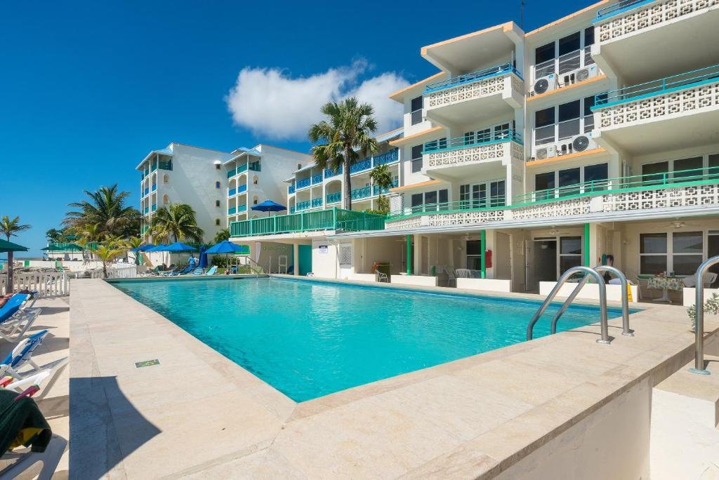 Área externa do Rostrevor Hotel. Uma piscina no centro, ao redor cadeiras de tomar sol e guarda-sóis. No fundo as varandas dos quartos. Imagem ilustrando post Hotéis em Barbados.