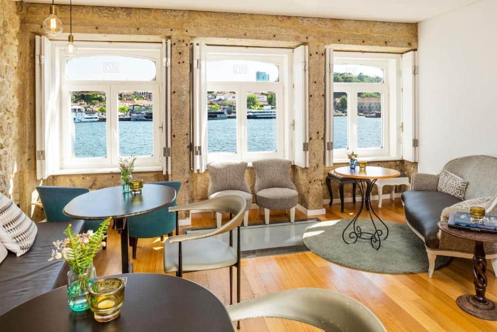 Sala de estar do 1872 River House com duas mesas redondas do lado esquerdo da imagem, com cadeiras, do lado direito uma poltrona cinca com uma mesa de madeira a frente. já na parte central ao fundo duas cadeiras estofadas a baixo da janelas.