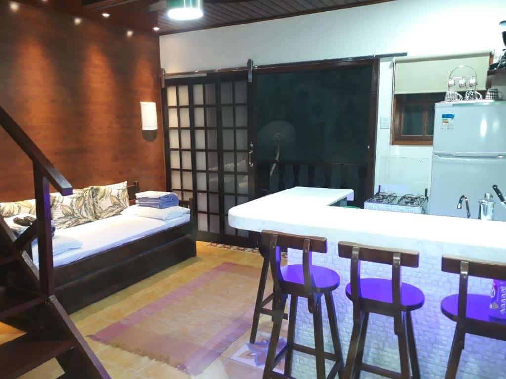 Sala e cozinha do airbnb Angel Ilha Flats e Varanda da Praia. No canto esquerdo há um sofá e é possível ver um pedaço da escada e já no canto direito há uma bancada com três banquinhos e atrás a cozinha, com um fogão e uma geladeira encostados na parede ao fundo. No canto esquerdo há uma porta que dá acesso a varanda.