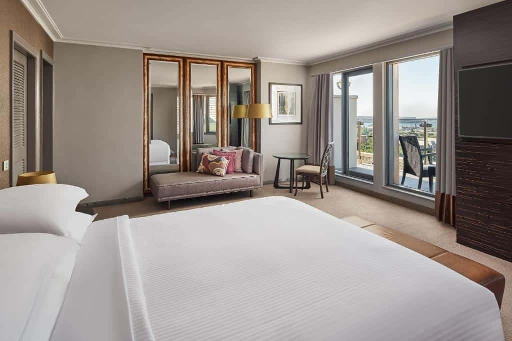 Quarto do Sheraton Grand Sydney Hyde Park. Uma cama de casal d lado esquerdo, no fundo um pequeno sofá, um armário, um abajur e uma mesa com cadeira. Do lado direito uma TV e a porta da varanda. 