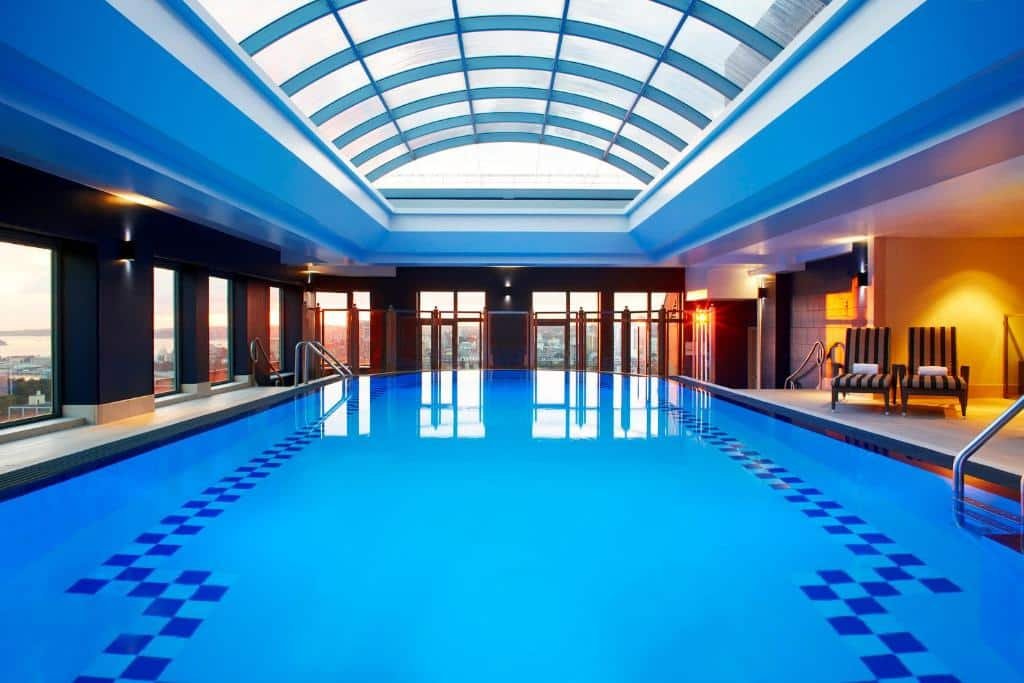 Piscina do Sheraton Grand Sydney Hyde Park. Uma piscina no meio coberta, do lado esquerdo e no fundo a paisagem da cidade. Do lado direito duas cadeiras de descanso.