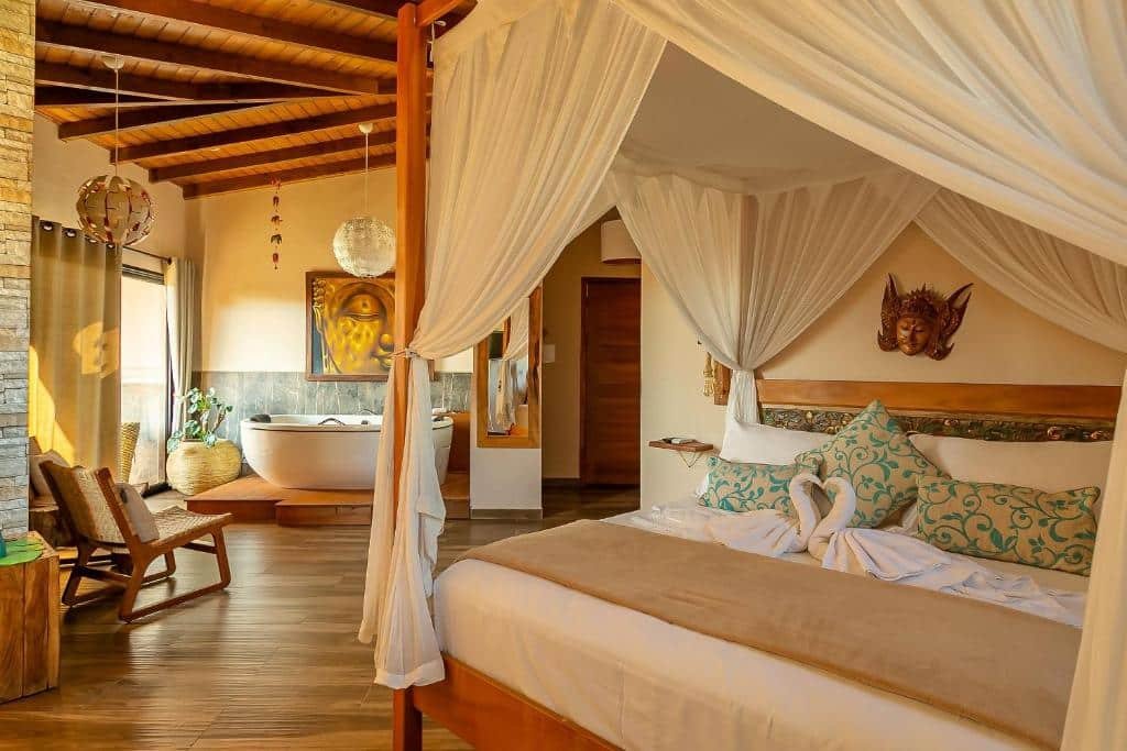 Quarto do hotel Shiva Boutique Hotel - Praia do Rosa. Uma cama de casal com dossel está do lado direito. Ao fundo, no lado esquerdo, há uma cadeira e uma banheira encostada na parede, abaixo de um quadro e ao lado de uma porta transparente com cortinas e um vaso de planta. No meio da imagem, entre a banheira e a cama, é possível ver um espelho na parede e uma porta fechada ao fundo.