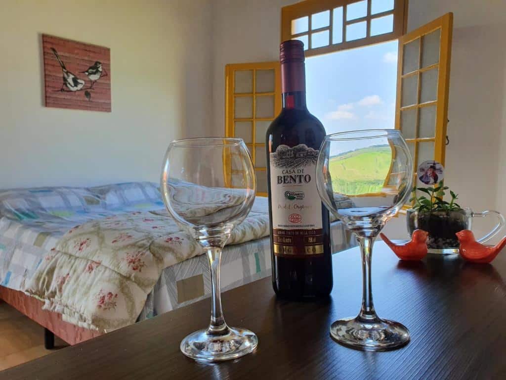Duas taças e uma garrafa de vinho em cima de um móvel de madeira, com uma cama ao fundo e uma janela com vista da natureza