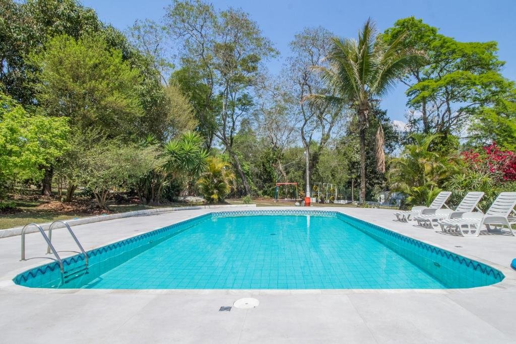 Piscina do Sitio só Alegria, em Guararema, com uma escadinha para entrar na água, 3 espreguiçadeiras brancas no lado direito, parquinho infantil ao fundo e bastante natureza em volta
