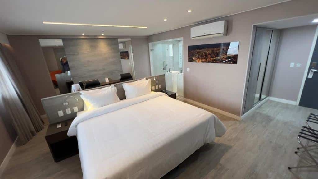 Grande quarto de hotel com cama de casal centralizada com colcha branca, mesa com duas cadeiras atrás da cama, porta de vidro para o banheiro ao lado, quadro decorativo e ar-condicionado na parede ao lado da cama. Imagem para ilustrar o post hotéis em Campina Grande