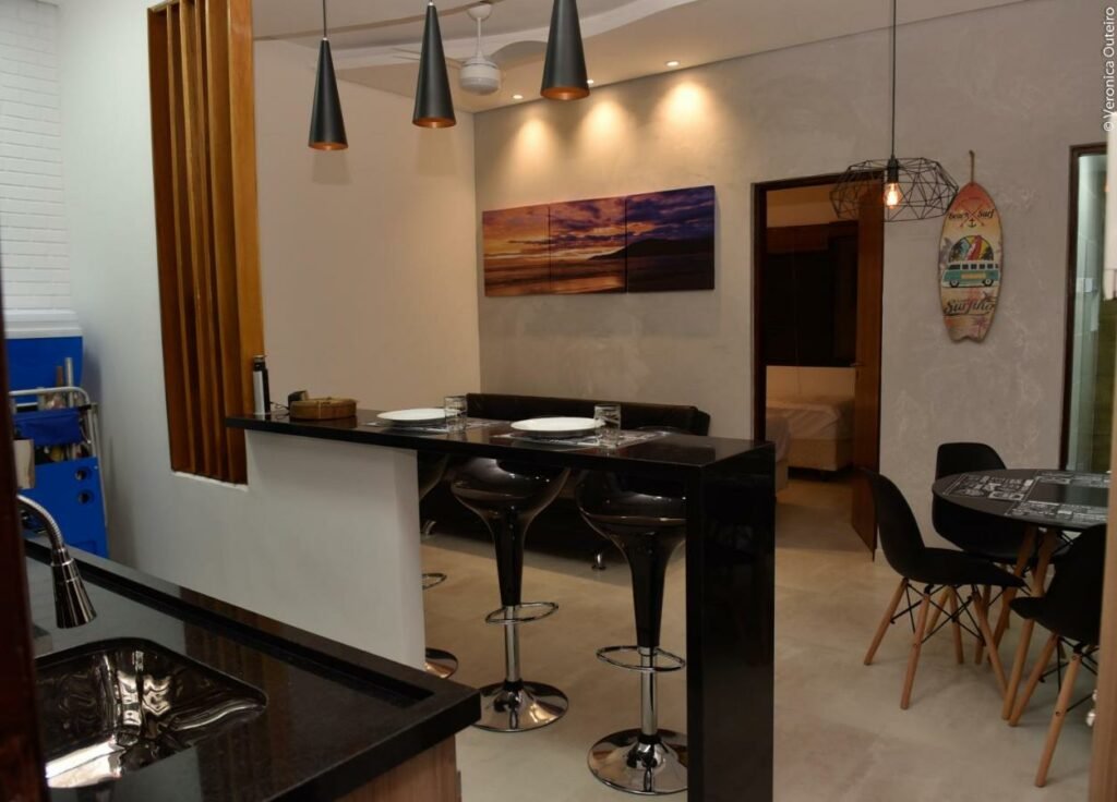 Área de estar e cozinha do Sun House Flat013 Residencial. Ao fundo, na área de estar, há um sofá ao lado de uma porta e uma mesa redonda de vários lugares. No meio do ambiente há um balcão servindo de mesa, com três bancos e alguns pratos em cima. Em primeiro plano é possível ver a pia da cozinha.