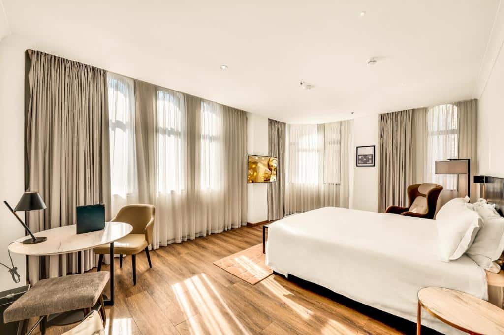 Quarto do The Grace Hotel. Uma cama de casal do lado direito, com uma poltrona e um abajur. Do lado esquerdo uma mesa com duas poltronas e uma TV. Três janelas no quarto. Imagem para ilustrar post sobre hotéis em Sydney.